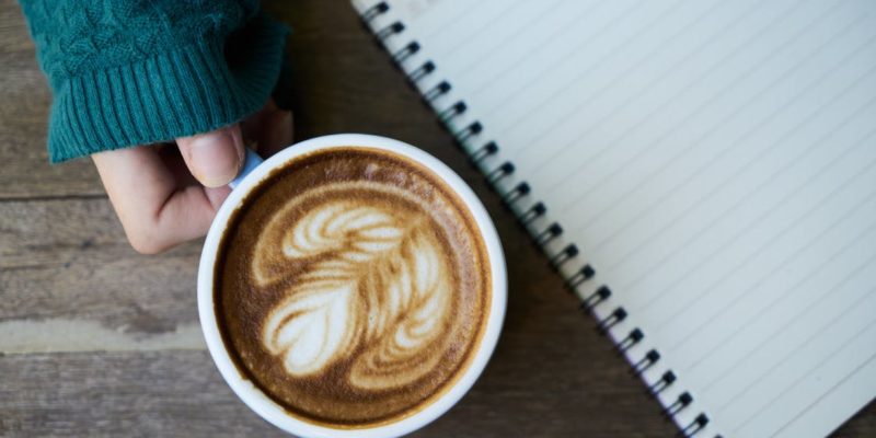 Person Holding White Ceramic Mug Beside Notebook