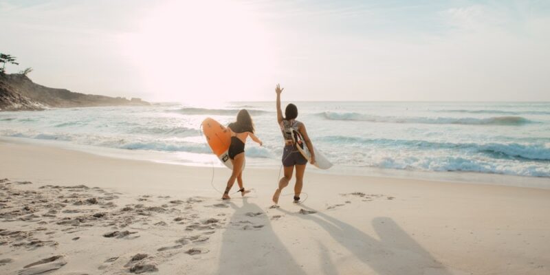 two women walking towards the ocean carrying surfboards during day